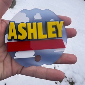 Peut inclure: Un panneau circulaire bleu et blanc avec une bande rouge et des nuages blancs. Le nom "ASHLEY" est &eacute;crit en lettres jaunes avec un contour bleu.