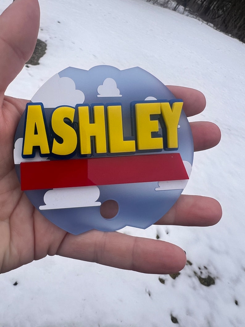 Peut inclure: Un panneau circulaire bleu et blanc avec une bande rouge et des nuages blancs. Le nom "ASHLEY" est &eacute;crit en lettres jaunes avec un contour bleu.