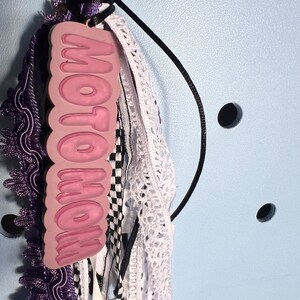 May include: A pink charm with the word "MOTO MOM" in white lettering, attached to a black cord with purple, white, and black ribbons.