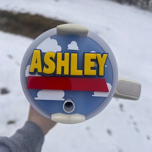 Peut inclure: Un couvercle rond bleu et blanc avec une bande rouge et un nom jaune "ASHLEY" dessus. Le couvercle a un trou pour paille et un bord blanc.