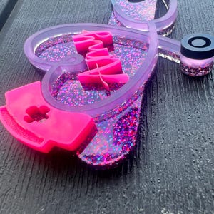 Pu&ograve; includere: Uno stetoscopio in resina viola e rosa con glitter incorporati nella resina. Lo stetoscopio ha la forma di un cuore con la parola "Infermiera" scritta in rosa all'interno del cuore. Un berretto da infermiera rosa &egrave; attaccato alla parte inferiore dello stetoscopio.