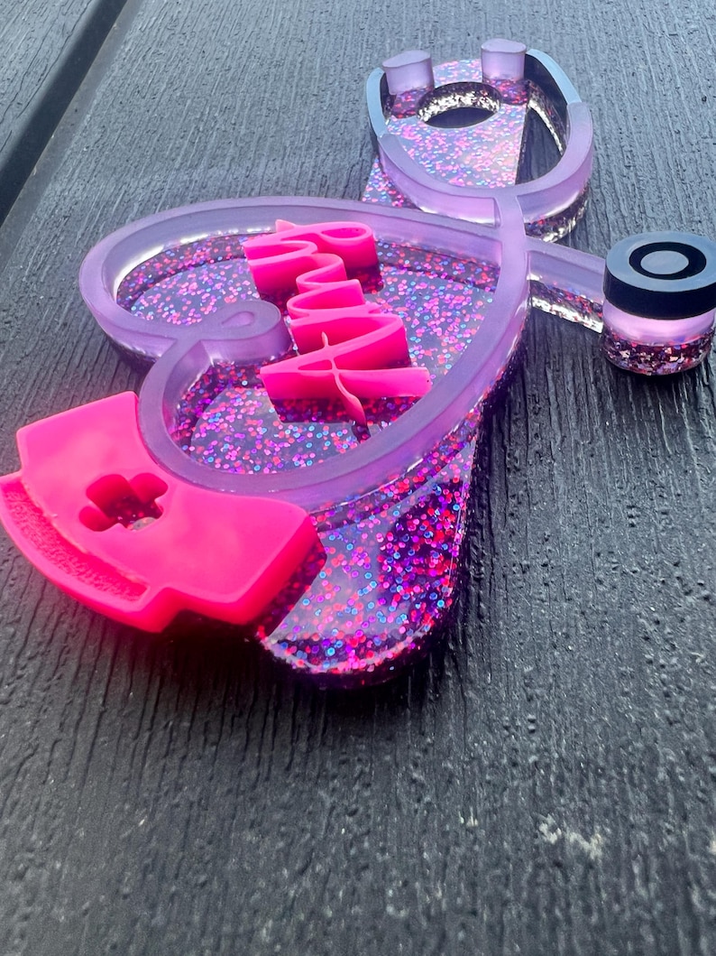 Pu&ograve; includere: Uno stetoscopio in resina viola e rosa con glitter incorporati nella resina. Lo stetoscopio ha la forma di un cuore con la parola "Infermiera" scritta in rosa all'interno del cuore. Un berretto da infermiera rosa &egrave; attaccato alla parte inferiore dello stetoscopio.