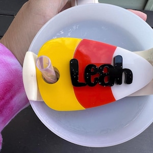 May include: A yellow, red and white candy cane-shaped lid with the name "Leah" in black glitter letters. The lid is designed to fit on a cup with a straw.