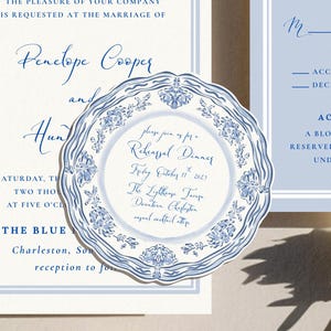 May include: A wedding invitation suite featuring a cream-colored main card, a light blue RSVP card, and a round rehearsal dinner card with a blue and white floral border. The text includes details for the marriage of Penelope Cooper and Hunter.
