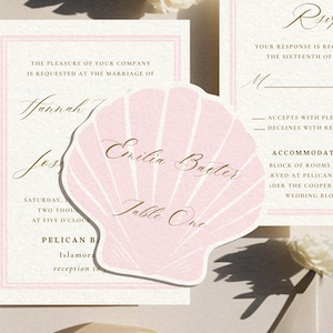 May include: A set of wedding stationery with a light pink theme. The set includes an invitation, RSVP card, and a seashell-shaped place card with gold script. The place card reads "Emilia Bacter" and "Table One".