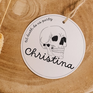 May include: White round tag with black text and a skull illustration. The text reads "til death do us party" and "Christina".