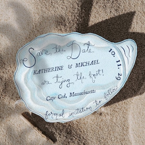 以下が含まれることがあります： 砂の上に置かれた、薄い青色の貝殻型の結婚式の招待状。テキストには「Save the Date」と書かれており、キャサリンとマイケルの結婚式が2029年10月11日にマサチューセッツ州ケープコッドで行われることを発表しています。「formal invitation to follow」と書かれています。