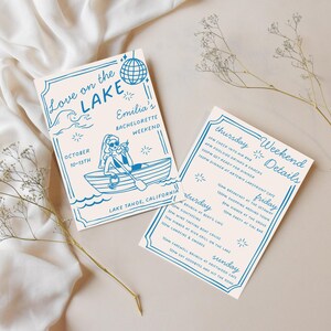 Puede incluir: Dos tarjetas de invitaci&oacute;n con texto e ilustraciones azules sobre un fondo crema. Una tarjeta dice "Love on the Lake" y "Emilia's Bachelorette Weekend". La otra tarjeta enumera los detalles del fin de semana. Flores secas y tela en el fondo.