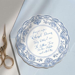 May include: A round, scalloped-edge paper invitation featuring a blue and white floral pattern around the border. The text reads: please join us for a Rehearsal Dinner, Friday, October 11, 2023, The Lighthouse Tavern, Downtown Charleston, casual cocktail attire.