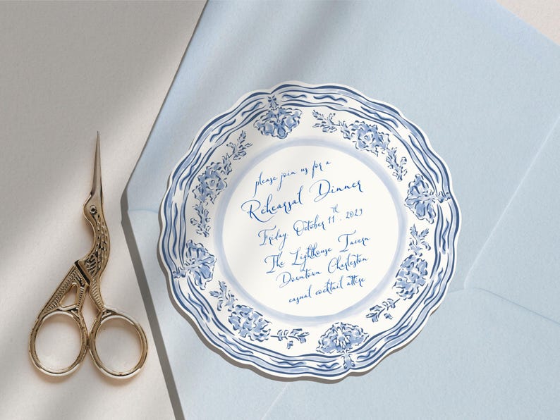 May include: A round, scalloped-edge paper invitation featuring a blue and white floral pattern around the border. The text reads: please join us for a Rehearsal Dinner, Friday, October 11, 2023, The Lighthouse Tavern, Downtown Charleston, casual cocktail attire.