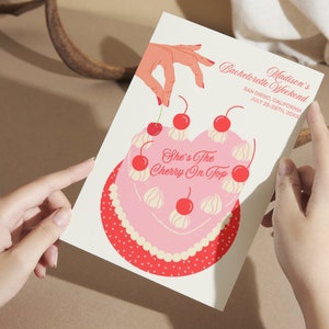 May include: A pink and red bachelorette party invitation with a heart-shaped cake decorated with white frosting and red cherries. The text on the invitation reads "Madison's Bachelorette Weekend, San Diego, California, July 25-28th, 20XX, She's The Cherry On Top."