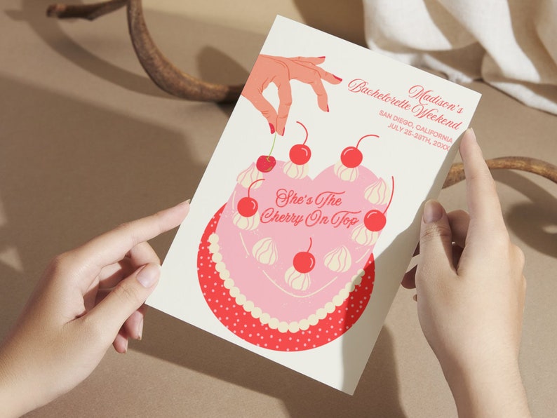 May include: A pink and red bachelorette party invitation with a heart-shaped cake decorated with white frosting and red cherries. The text on the invitation reads "Madison's Bachelorette Weekend, San Diego, California, July 25-28th, 20XX, She's The Cherry On Top."