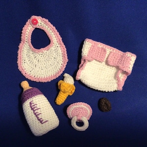 May include: A collection of handmade crochet baby items, including a bib, diaper, bottle, banana, teether, and a small brown object. The items are primarily white with pink and purple accents, set against a blue background.