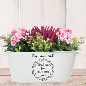 May include: White metal planter with a black inscription that reads "Mrs. Hammond Thank You for Helping Me Grow" surrounded by a wreath design. The planter is filled with pink and purple flowers.