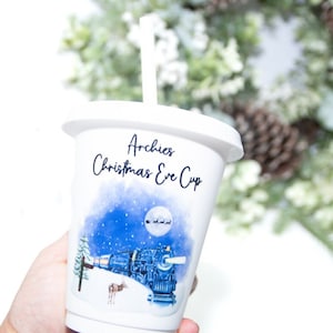 Op de afbeelding: Witte plastic beker met een blauw en wit kerstontwerp met een trein, een hert en een sterrenhemel. De beker heeft een rietje en staat "Archie's Christmas Eve Cup" in zwarte tekst.