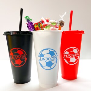 May include: Three personalized cups with soccer ball designs and names "Lewis", "Jaxon", and "Harry". The cups are black, white, and red. The white cup is filled with colorful candies.