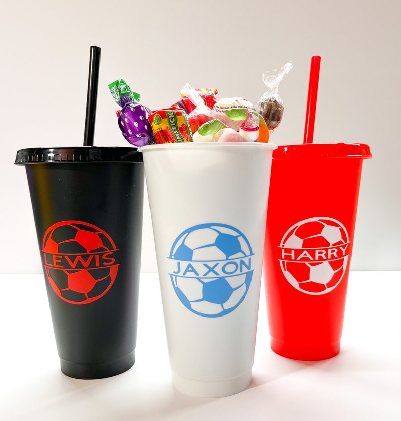 May include: Three personalized cups with soccer ball designs and names "Lewis", "Jaxon", and "Harry". The cups are black, white, and red. The white cup is filled with colorful candies.
