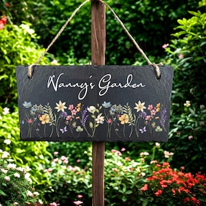 Könnte beinhalten: Rechteckiges schwarzes Schiefer-Gartenschild mit dem weißen Schriftzug "Nanny's Garden". Der untere Rand zeigt ein farbenfrohes Blumendesign mit verschiedenen Blumen und Schmetterlingen. Das Schild hängt an einem Holzpfosten mit Schnur.