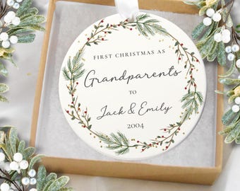 Peut inclure: Ornement de Noël rond en céramique blanche avec une couronne botanique. Le texte indique « Premier Noël en tant que grands-parents de Jack & Emily 2004 ». Il est suspendu à un ruban blanc et se trouve dans un coffret cadeau marron.