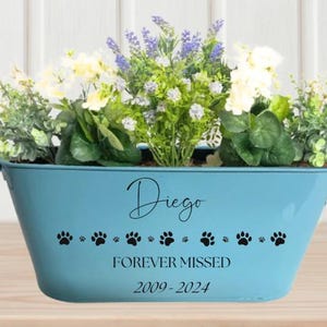 May include: A light blue metal planter with the name "Diego" and the text "Forever Missed 2009-2024" in black lettering. The planter is filled with white and purple flowers and has a row of black paw prints along the bottom.