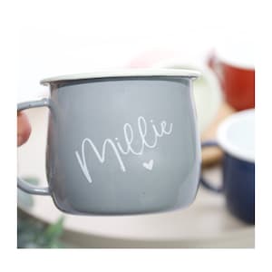 K&ouml;nnte beinhalten: Eine graue Emaille-Tasse mit wei&szlig;em Rand und einer wei&szlig;en Aufschrift "Millie" mit einem kleinen Herz.