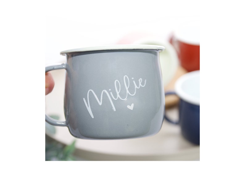 K&ouml;nnte beinhalten: Eine graue Emaille-Tasse mit wei&szlig;em Rand und einer wei&szlig;en Aufschrift "Millie" mit einem kleinen Herz.