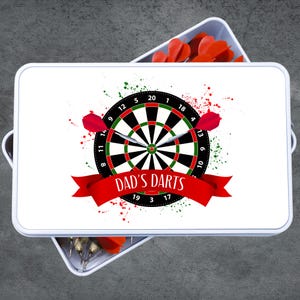 May include: A white rectangular tray with a dartboard design. The dartboard is black, green, and red, with the text "DAD'S DARTS" on a red banner. Two red darts are positioned on the board, and the tray is filled with red dart flights.