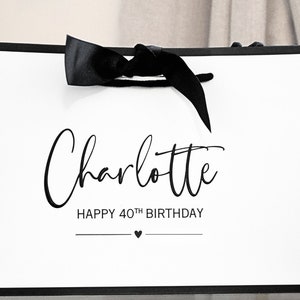 May include: White gift bag with a black ribbon and black text that says "Charlotte Happy 40th Birthday" with a small black heart.