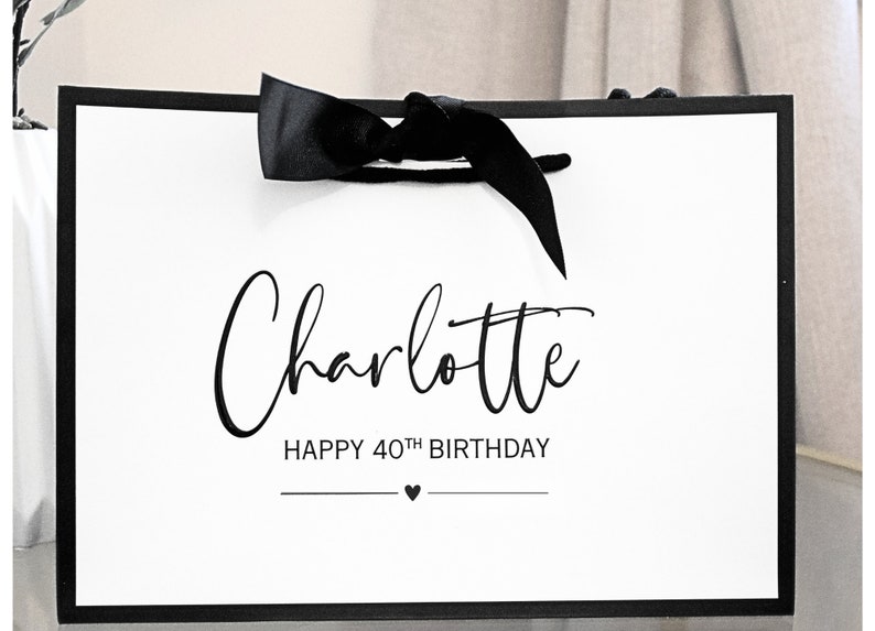 May include: White gift bag with a black ribbon and black text that says "Charlotte Happy 40th Birthday" with a small black heart.