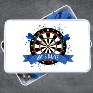 May include: A white rectangular box with a dartboard design. The dartboard is black, red, and green with blue darts. The text "DAD'S DARTS" is on a blue banner. Additional blue darts are visible.
