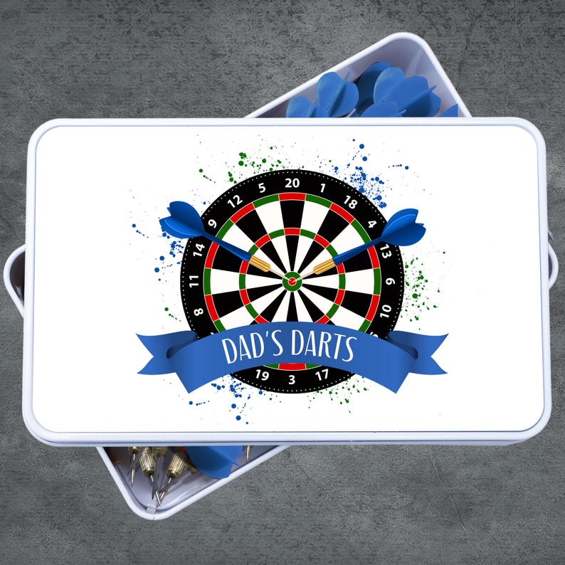 May include: A white rectangular box with a dartboard design. The dartboard is black, red, and green with blue darts. The text "DAD'S DARTS" is on a blue banner. Additional blue darts are visible.
