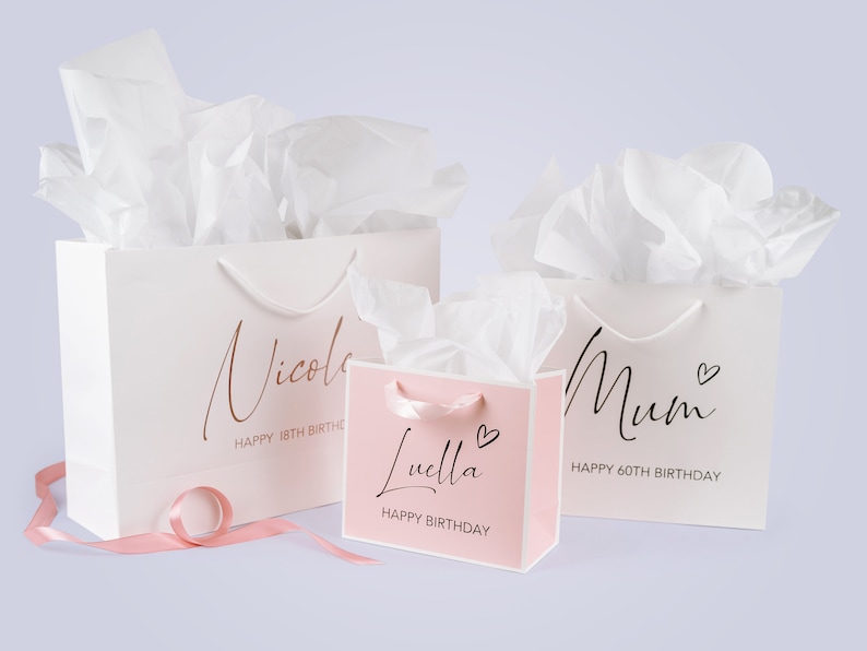Peut inclure: Trois sacs cadeaux blancs avec des accents roses et du papier de soie. Les sacs sont personnalis&eacute;s avec les noms "Nicole", "Luella" et "Maman" et comprennent des salutations d'anniversaire.
