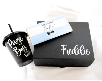 May include: A black gift box with the name "Freddie" written in white on the top. The box contains a chocolate bar with the text "Will you be our Page Boy?" and a black cup with a straw that says "Page Boy" with the name "Freddie" written underneath.