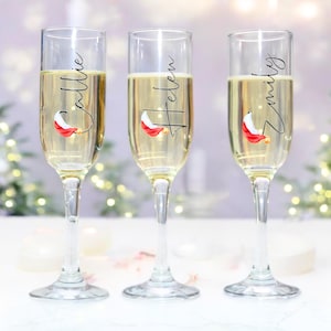 May include: Three clear glass champagne flutes with personalised names "Callie", "Helen", and "Emily" written in black cursive. Each flute has a small red and white Santa hat graphic.