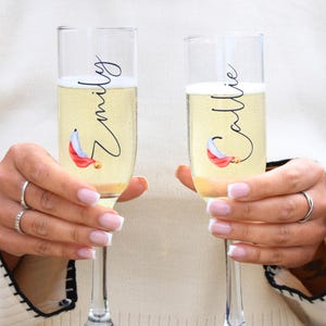 May include: Two clear glass champagne flutes with personalized names "Emily" and "Callie" in black script. Each flute features a festive red and white Santa hat graphic.