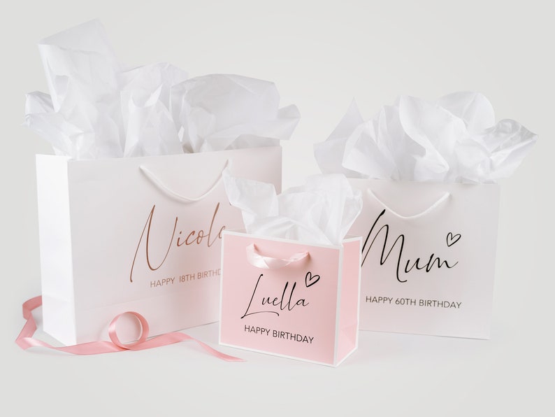 Peut inclure: Trois sacs cadeaux blancs avec des accents roses et du papier de soie. Les sacs sont personnalis&eacute;s avec les noms "Nicole", "Maman" et "Luella" et le texte "Joyeux 18&egrave;me anniversaire", "Joyeux 60&egrave;me anniversaire" et "Joyeux anniversaire".