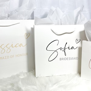 May include: Three white gift bags with handles. The bags are decorated with gold, black, and pink lettering. The text on the bags reads "Jessica Maid of Honour", "Sofia Bridesmaid", and "Callie Flower Girl".