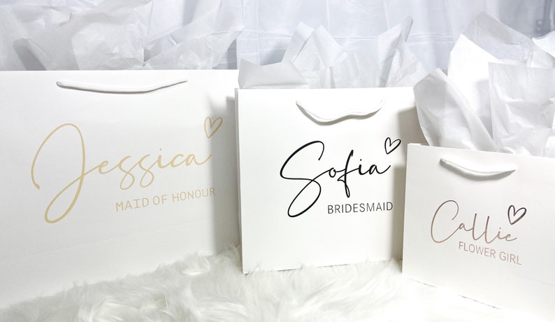 May include: Three white gift bags with handles. The bags are decorated with gold, black, and pink lettering. The text on the bags reads "Jessica Maid of Honour", "Sofia Bridesmaid", and "Callie Flower Girl".
