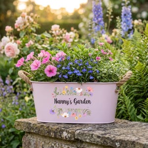 May include: A pale pink metal planter filled with pink and blue flowers and green foliage. The planter has rope handles and the words "Nanny's Garden" in a floral wreath design.