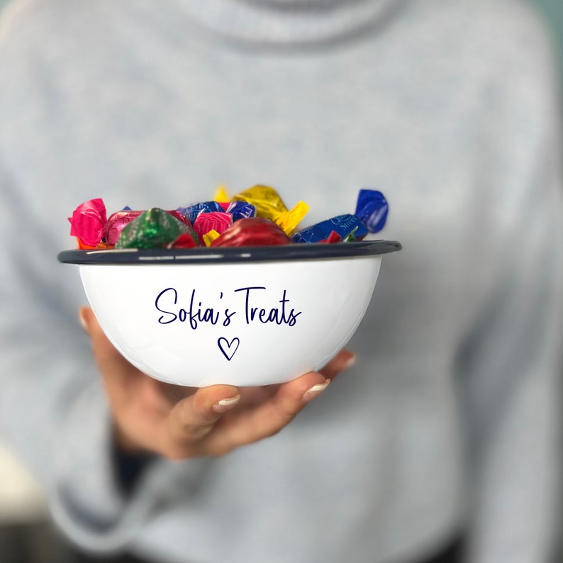 May include: A white enamel bowl with a navy blue rim, filled with colourful wrapped sweets. The bowl has the text "Sofia's Treats" and a heart symbol. The bowl is being held by a person.