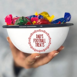 Pode incluir: Tigela de esmalte branco com borda azul cheia de doces coloridos embrulhados. A tigela tem um design circular vermelho com o texto "Dad's Football Treats" no centro.