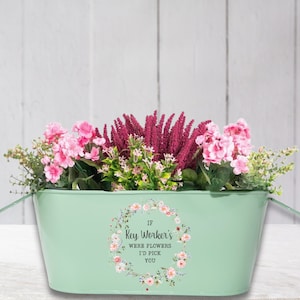 Könnte beinhalten: Ein mintgrüner Metallpflanztopf gefüllt mit rosa und burgunderfarbenen Blumen und Grün. Der Pflanztopf hat ein Blumengirlanden-Design mit dem Text "If Key Worker's Were Flowers I'd Pick You". Der Pflanztopf hat zwei Griffe.