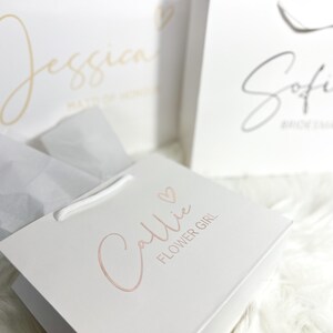 May include: Three white gift bags with pink and gold lettering. The bags are labeled "Jessica Maid of Honor", "Sofia Bridesmaid", and "Callie Flower Girl".