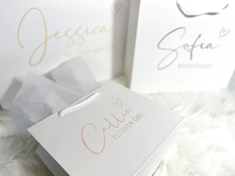 May include: Three white gift bags with pink and gold lettering. The bags are labeled "Jessica Maid of Honor", "Sofia Bridesmaid", and "Callie Flower Girl".