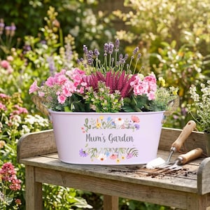 May include: A lilac metal planter filled with a variety of colourful flowers and greenery. The planter has rope handles and the words "Mum's Garden" are printed on the front. A small trowel and hand rake rest on a wooden bench.