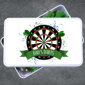 May include: White rectangular box with a dartboard design. The dartboard is black, red, and white with green darts. The text "DAD'S DARTS" is on a green banner. Blue and green paint splatters are in the background.