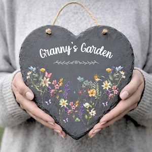 Può includere: Cartello da giardino a forma di cuore in ardesia con la scritta "Granny's Garden" in bianco. Il cartello presenta un colorato disegno floreale con farfalle e un uccellino. Una corda di iuta è attaccata per appenderlo.