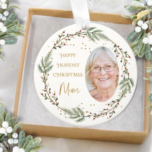 Könnte beinhalten: Weißer Keramik-Weihnachtsschmuck mit einem Foto einer Frau, umgeben von einem Kranz aus grünen Blättern und roten Beeren. Der Schmuck trägt die Worte "Happy Heavenly Christmas Mum" in goldener Schrift und wird in einer braunen Schachtel präsentiert.
