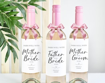 May include: Three wine bottles with labels that say "Pairs well with Mother of the Bride", "Pairs well with Father of the Bride", and "Pairs well with Mother of the Groom". The bottles are tied with pink ribbon bows.