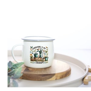 Puede incluir: Taza de esmalte blanco con borde gris. La taza tiene una ilustración colorida de una escena de jardín con un pozo, una regadera y herramientas de jardinería. El texto "GARDENING IS MY THERAPY" está impreso en la taza.